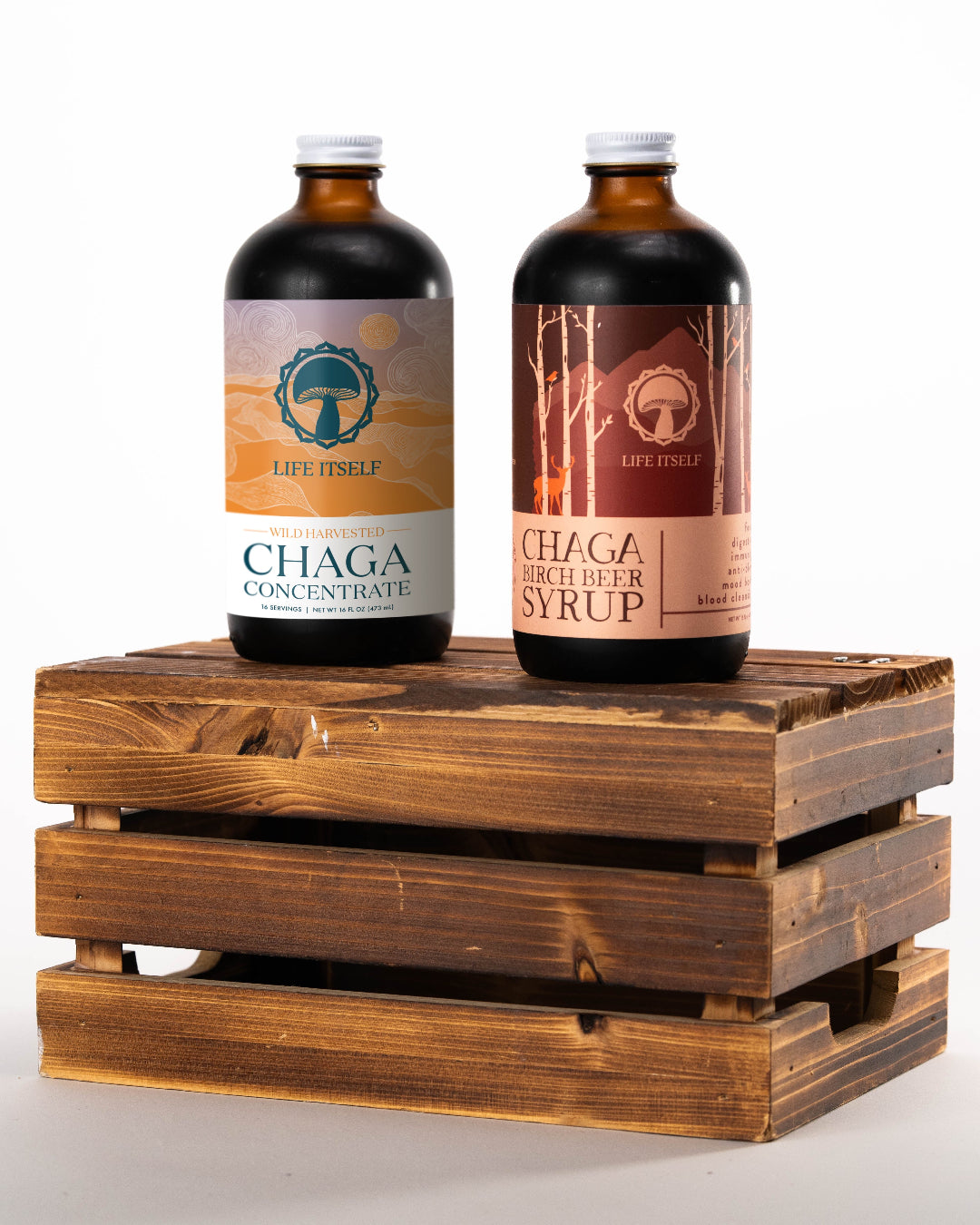 Two bottles of Life Itself mushroom products resting on a rustic wooden crate against a white background, featuring Wild Harvested Chaga Concentrate and Chaga Birch Beer Syrup.